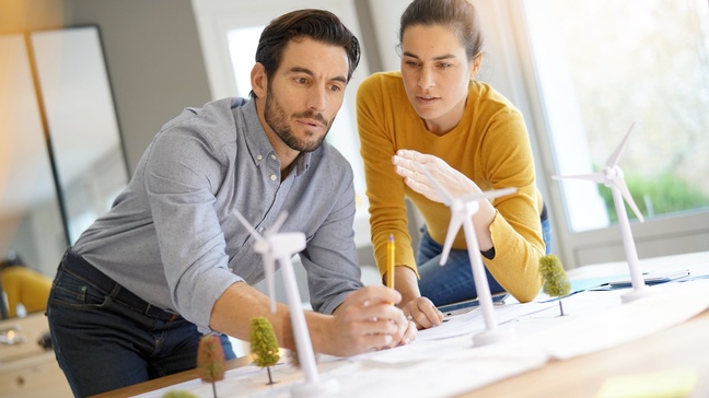 Zwei Personen beugen sich über eine Tischplatte. Auf der Tischplatte ist ein großes, weißes Papier ausgebreitet. Darauf stehen Miniaturbäume sowie Miniaturwindräder. Beide Personen blicken auf die Modelle vor ihnen.
