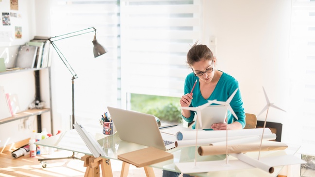 Person mit Brille und geschlossenen Haaren sitzt an einem Schreibtisch mit Windrad-Modellen, einem Laptop und blickt auf ein Tablet während ein Stift in der Hand gehalten wird, Home Office