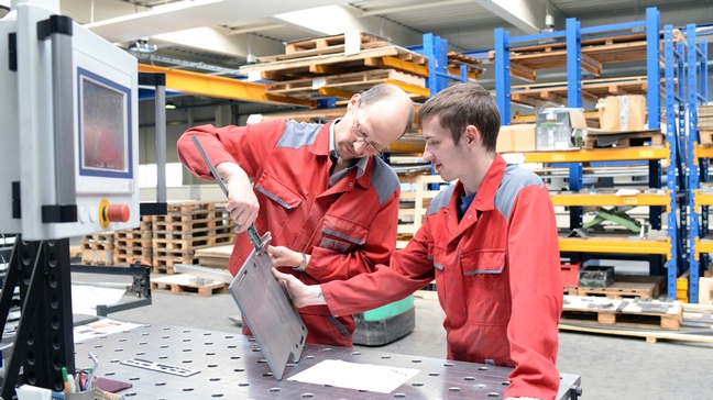 Zwei Personen in roten Arbeitsoveralls an Arbeitsplatte in Werkhalle vermessen Ösen eines Metallstückes
