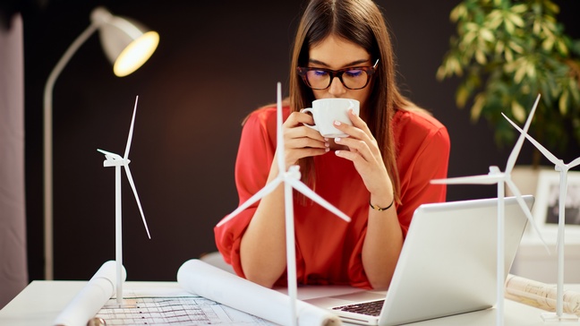 Person mit Brillen sitzt an Schreibtisch und nippt an Kaffee, ringsum am Tisch Modelle von Windrädern, Pläne und aufgeklappter Laptop