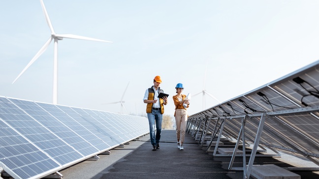 Zwei Personen mit Schutzhelmen spazieren durch Anlage mit Solarpaneelen, im Hintergrund Windräder