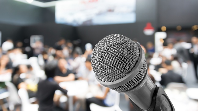 Nahaufnahme eines Mikrofons vor einer Halle, in der Personen in Gruppen an Tischen sitzen