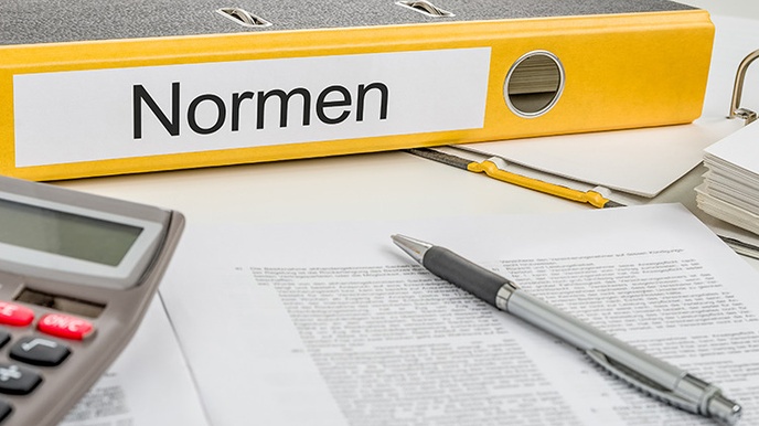 Gelber Ordner mit Beschriftung Normen auf Längsseite liegend, davor Stift auf Dokument und Ausschnitt eines Taschenrechners