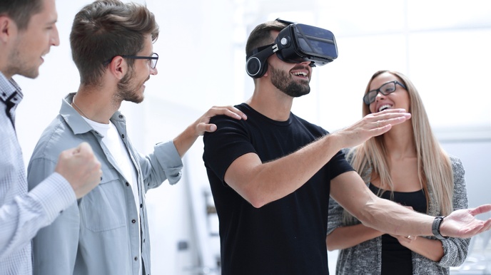 Eine Person trägt eine Virtual Reality Brille am Kopf und greift mit den Armen während andere Personen daneben stehen und freudig zusehen