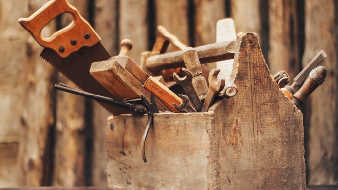 In einer Werkzeugkiste aus Holz sind verschiedene Werkzeuge, zum Beispiel unterschiedliche Zangen, ein Hobel, ein Hammer und eine Säge.