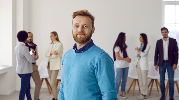 Person mit rötlichem Haar und Bart sowie blauem Pullover blickt freudig in die Kamera, im Hintergrund stehen weitere Personen und unterhalten sich in der Unschärfe