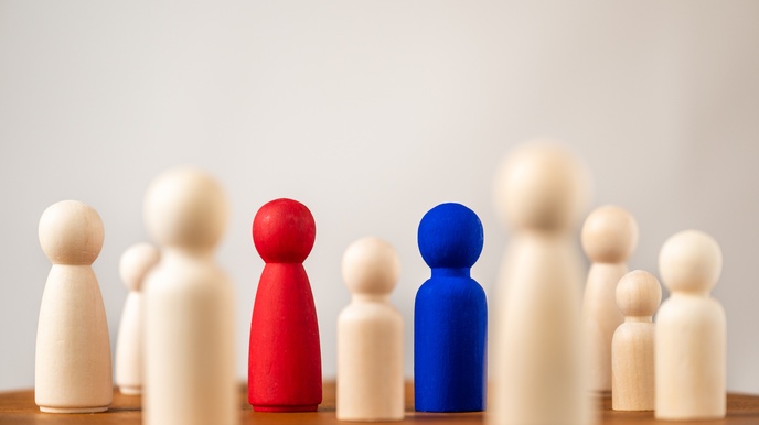 Aufgestellte Spielfiguren aus Holz, von denen 2 rot und blau sowie im Fokus sind. Sie stehen auf einem braunen Holztisch