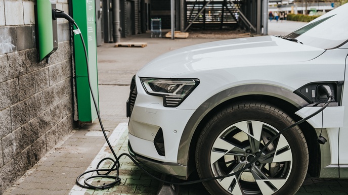 Ein weißes Elektroauto ist zur Hälfte zu sehen. Es wird an einer E-Tankstelle aufgeladen, die durch ihre grüne Farbe das Bild dominiert. Die Ladestation befindet sich an einer Steinmauer.  Im Hintergrund sieht man verschwommen eine  Garage, 