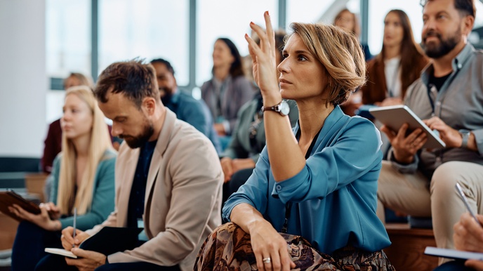 In Hörsaal sitzende Person in Seitenansicht im Fokus, die Hand in die Höhe hebt, im Hintergrund ringsum verschwommen weitere sitzende Personen