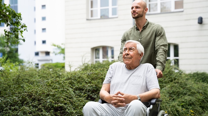 Eine Person in einem Hemd schiebt eine andere Person in einem Rollstuhl durch eine Parkanlage. Im Hintergrund sind Gebäude. Links neben ihnen sind grüne Büsche