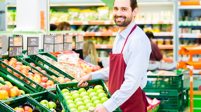 Seitliches Halbporträt einer Person mit roter Schürze, die mit beiden Händen eine niedrige, grüne Kiste hält, in der grünes Obst liegt. Links sind weitere dieser Kisten, befüllt mit unterschiedlichem Obst. Im Hintergrund sind Regale mit Lebensmitteln