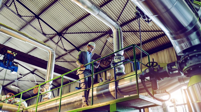 Person in Businesskleidung und Schutzhelm steht auf einer Plattform und blickt auf ein Tablet in einem Innenraum eines Kraftwerkes