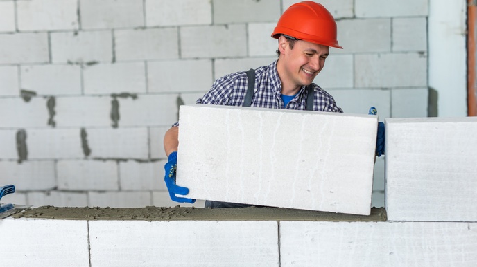 Person mit kariertem Hemd, Anzug, rotem Helm und Schutzhandschuhen baut eine Ziegelwand mit grauen Silikatziegeln