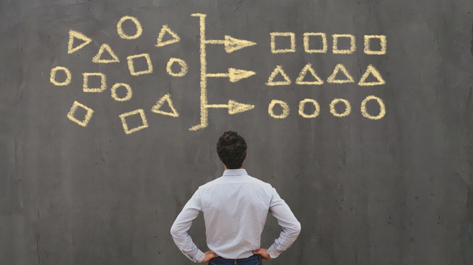 Eine Person ist von hinten fotografiert und steht vor einer schwarzen Tafel. Darauf sind mit gelber Kreide links Rechtecke, Dreiecke und Kreise gezeichnet. In der Mitte ist ein Strich, von dem drei Pfeile ausgehen, die Symbole sind rechts in einer Reihe