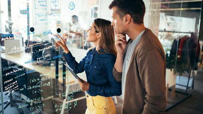 Zwei Personen stehen vor transparenter Wand, auf der Formeln und Schriften abgebildet sind, hinter der Wand verschwommen Großraumbüro