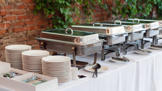 Auf einem Tisch mit weißem Tischtuch stehen gestapelte Teller, Besteck und fünf silberne Speisenwärmer nebeneinander. Vor jedem Behälter liegt eine Zange oder ein größer Löffel auf einem Teller.