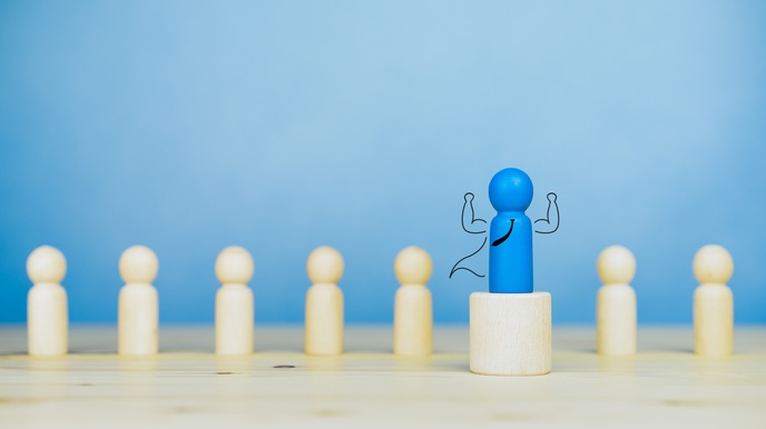 Kleine blaue Holzfigur steht auf Holzpodest, illustriert Krawatte, Umhang und erhobene starke Arme, im Hintergrund weitere naturfarbene Holzfiguren