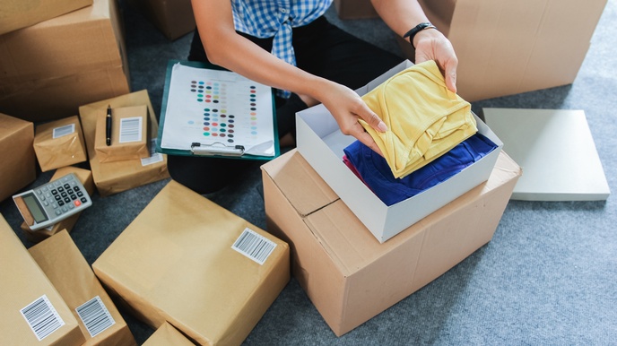 Mehrere, unterschiedlich große Kartons stehen auf dem Boden. Ein Karton ist geöffnet. Die Hände einer Person legen in diesen Karton ein gelbes Kleidungsstück. Darunter sind weitere Kleidungsstücke.