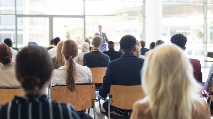 Rückenansicht vieler Menschen, die in Sesselreihen in einem großen Raum mit Fensterwänden sitzen. Im Hintergrund steht eine Person an einem Rednerpult, die zum Publikum spricht