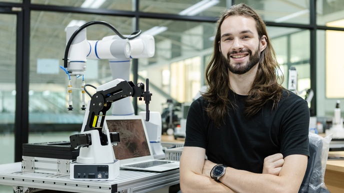 Lächelnde Person mit Bart und langem Haar im Portrait hält Arme vor Körper verschränkt, im Hintergrund Tisch mit aufgeklapptem Notebook und Roboterarm