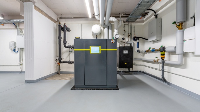 Ansicht einer Wärmepumpe: Dunkelgrauer metallischer Block mit gelber Linie und mittig platziertem Display in weißem Keller, ringsum Leitungen und Rohre verlaufend