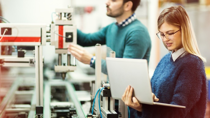 Person mit Brille in einer technischen Produktion hält einen Laptop in der Hand und blickt darauf, zweite Person in Unschärfe bedient eine Maschine im Hintergrund