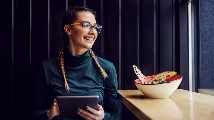 Lächelnde Person mit Brillen hält Tablet in Händen und blickt aus Fenster, neben ihr auf Sims Schale mit Brot und Salat stehend