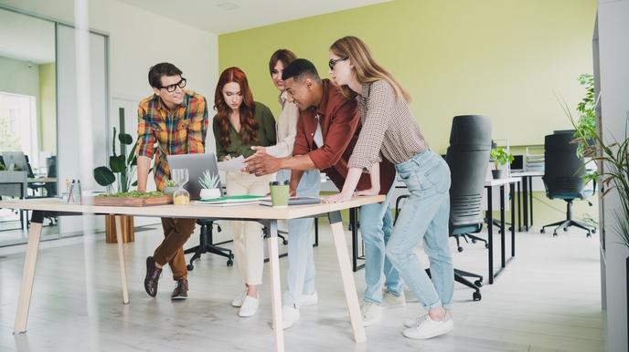 In einem Büro stehen um einen Tisch fünf Personen. Auf dem Tisch ist ein Laptop, auf den zwei Personen deuten. Eine dritte Person notiert etwas auf einem Zettel. Im Hintergrund sind Schreibtische und Sessel