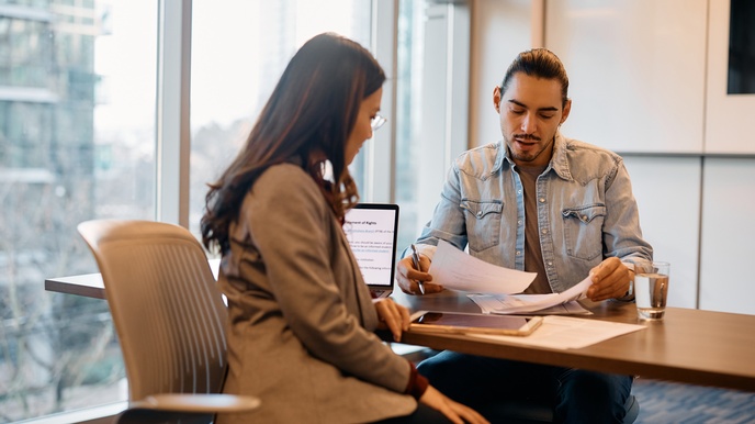 Zwei Personen sitzen sich an einem Tisch gegenüber. Vor der linken Person sind ein Laptop und Tablet. Die andere Person hält Zettel. Beide blicken auf die Zettel.