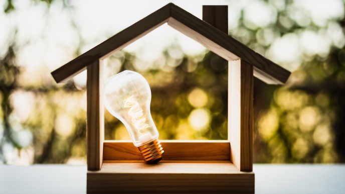 Miniaturholzhaus steht bei einem Fensterbrett und darin leuchtet eine  angelehnte Glühbirne, im Hintergrund zeigt sich ein grün, gelbes Bokeh