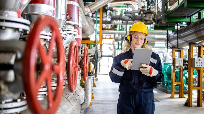 Eine Person mit gelbem Helm, Ohrenschützern, Arbeitskleidung, Handschuhen und Tablet, auf das sie blickt, steht in einem asphaltierten Gang in einem hohen Raum mit Rohren, Leitungen und Industrieanlagen. Direkt links neben ihr sind drei rote Radventile.