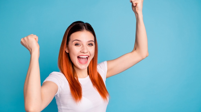 Eine junge Person mit rötlichen Haaren und einem lächelndem Mund steht vor einem blauem Hintergrund und hält beide Arme in die Höhe