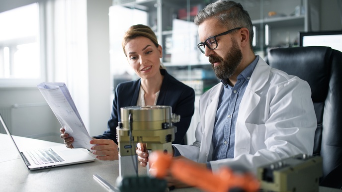 Zwei Personen sitzen an Arbeitstisch, eine Person hält Unterlagen in Händen, die andere hält Bauteil eines Roboterarms,  auf den beide Personen blicken