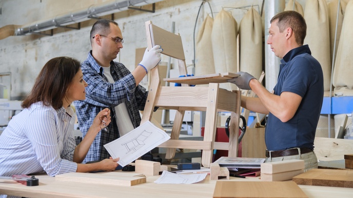 Seitliche Aufnahme dreier neben einem Tisch stehender Personen. Eine Person beugt sich über den Tisch und hält einen Zettel mit der Skizze eines Stuhls. Die anderen beiden Personen halten zwischen sich einen Holzstuhl.
