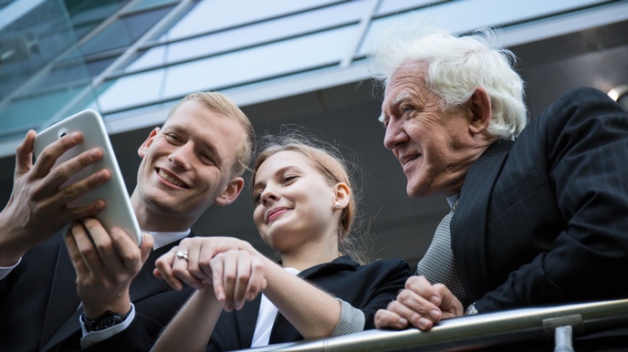 Drei Personen in dunklen Anzügen stehen dicht nebeneinander an einem Geländer und lehnen sich an. Die Person links hält ein Tablet in den Händen. Alle drei schauen in den Bildschirm
