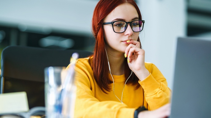 Person mit Brille in gelbem Pullover und gelben Fingernägeln sitzt vor aufgeklapptem Notebook mit Kopfhörern in den Ohren und fasst sich mit einer Hand an den Mund, im Vordergrund verschwommen Glas mit Stiften