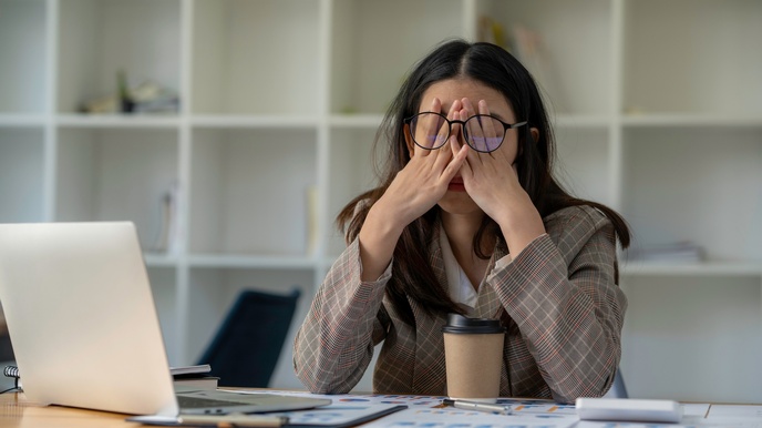 Eine Person sitzt an einem Tisch mit aufgeklappten Laptop, Unterlagen und einem Kaffeebecher. Sie hat die Hände auf ihr Gesicht gelegt