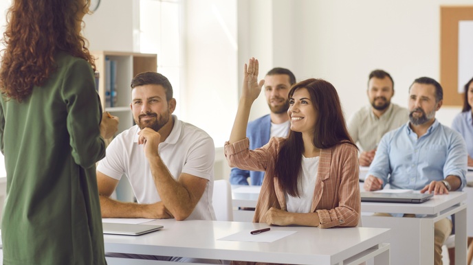 Frontalaufnahme von jeweils zwei Personen, die hinter einem Tisch sitzen. Im Bild sind insgesamt drei Tische und sechs Personen. Eine Person hebt die Hand. Alle blicken zu einer stehenden Person, die mit dem Rücken zur Kamera gewandt ist