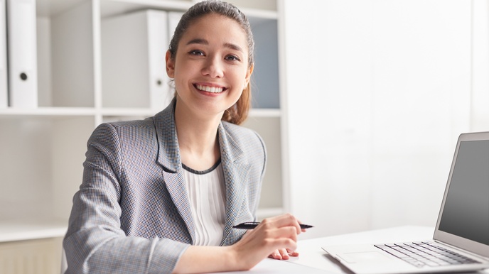 Porträt einer an einem Tisch sitzenden Tisch. Vor ihr liegt ein Clipboard. Sie hält einen Kugelschreiber in der rechten Hand. Schräg links vor ihr steht ein aufgeklappter Laptop. Hinter ihr ist ein weißes Regal
