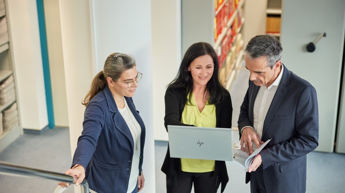 Drei Personen stehen in einem Flur, eine Person in der Mitte hält einen Laptop in Händen, eine Person neben ihr blickt gespannt darauf, während ein geöffnetes Buch in Händen gehalten wird