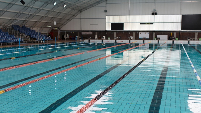 Ein leeres Hallenbad mit einem breiten und langen Pool, das durch rot-schwarze, schwimmende Linien in mehrere Bahnen unterteilt ist. Am linken Rad stehen mehrere blaue Sesselreihen