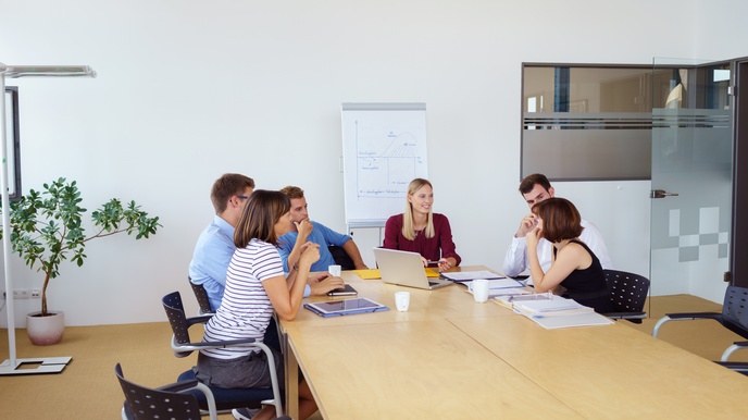 Personen sitzen rund um einen Besprechungstisch in einem Raum, vorne steht ein Flipchart mit einer aufgezeichneten Grafik