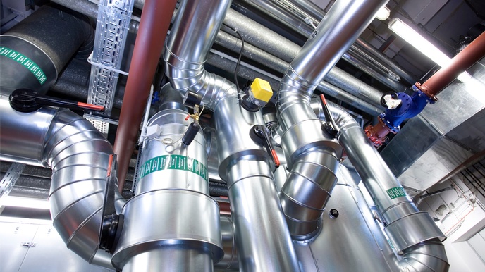 Dicke, metallische Rohre stehen in einem hohen Raum. Die Rohre machen an manchen Stelle eine Kurve und sind geschwungen. Die Rohre führen zu weiteren Rohren, die im Raum an der Decke oder sonstigen Stellen montiert sind