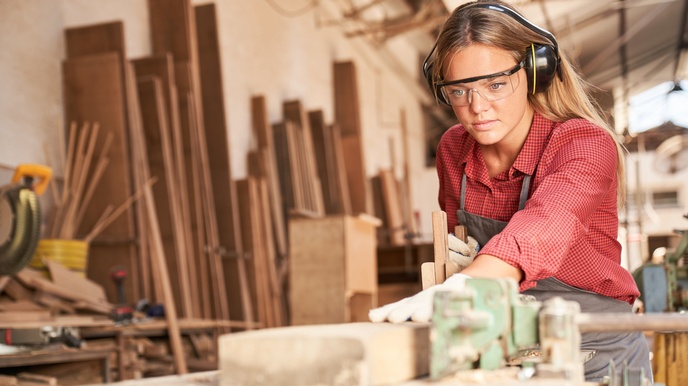 Person mit Ohrenschutz und Schutzbrillen hält Holzbalken auf Hobelbank, im Hintergrund verschwommen Holzbretter an Wand lehnend