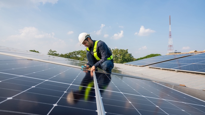 Auf einem leicht schrägen Dach sind viele dunkle Platten montiert. Die Platten werden von einem Metallrahmen umfasst. Auf den Platten ist eine Person mit Helm. Sie hält einen Bohrer in der Hand. Der Himmel ist blau und leicht bewölkt