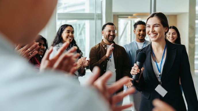 Personen mit Ausweisschildern stehen in einem Halbkreis und klatschen. Im Kreis steht eine Person mit dunklem Sakko, Mikrofon sowie Ausweisschild, die lächelt