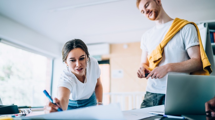 Person streckt sich über Tisch und markiert etwas mit Stift auf großem Papierbogen, andere Person sieht ihr mit geneigtem Kopf zu