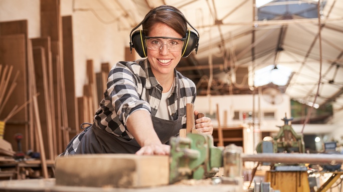 Person mit Ohrenschutz und Schutzbrillen hält Holzbrett in Tischkreissäge, nebenstehend weitere Person mit Ohrenschutz und Schutzbrillen Bretter haltend,  im Hintergrund verschwommen Holzbretter an Wand lehnend