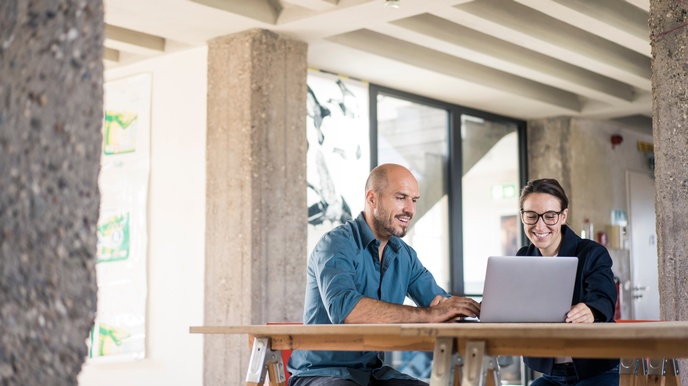 zwei Personen sitzen an einem Holztisch und schauen auf den Bildschirm eines Laptops