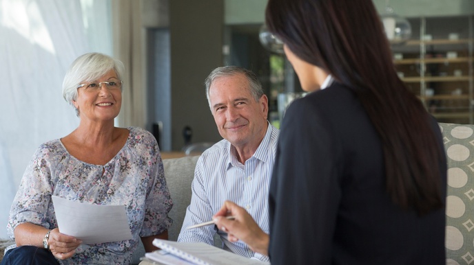 Rückenansicht einer sitzenden Person mit offenen, dunklen Haaren, die ein Heft auf ihrem Schoß hat. Ihr gegenüber sitzen zwei Personen mit grauen Haaren, die zu der anderen Person blicken. Die linke der beiden Personen hält einen Zettel